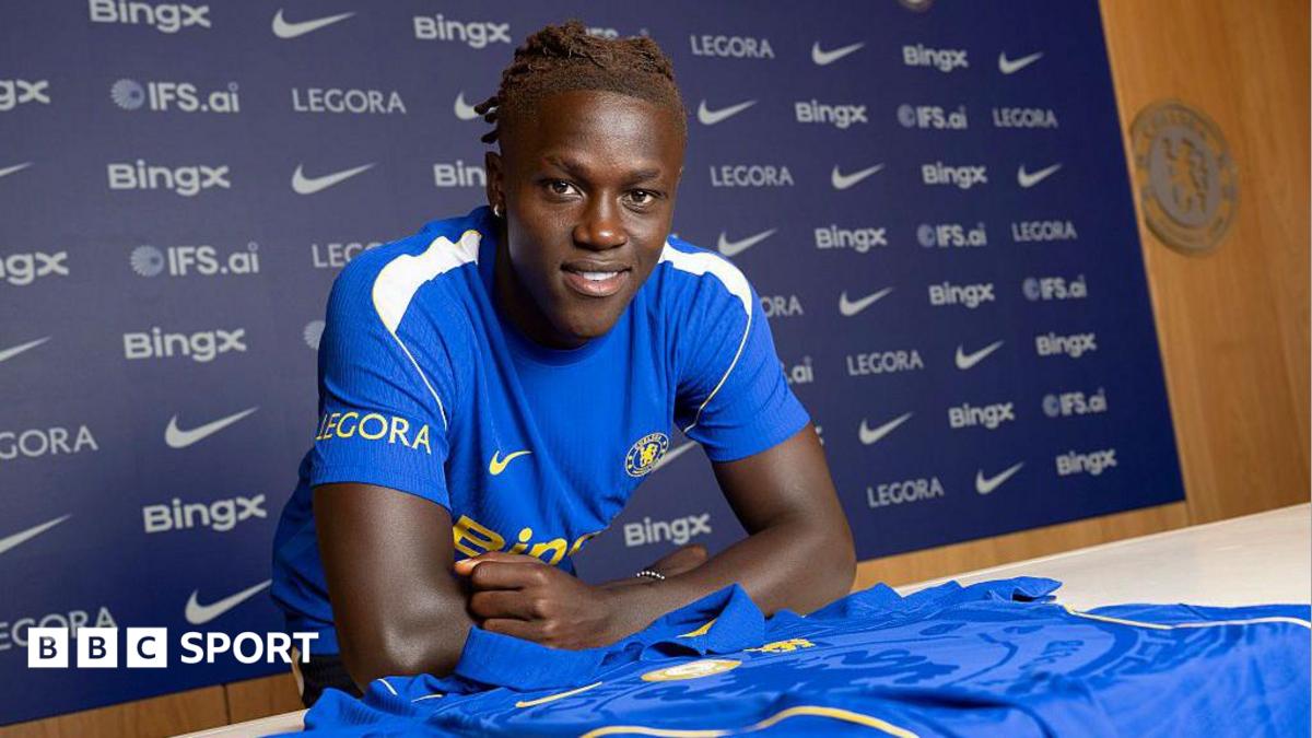 Quenda resting his arms on a table with a Chelsea shirt on and in front of him after signing for the club
