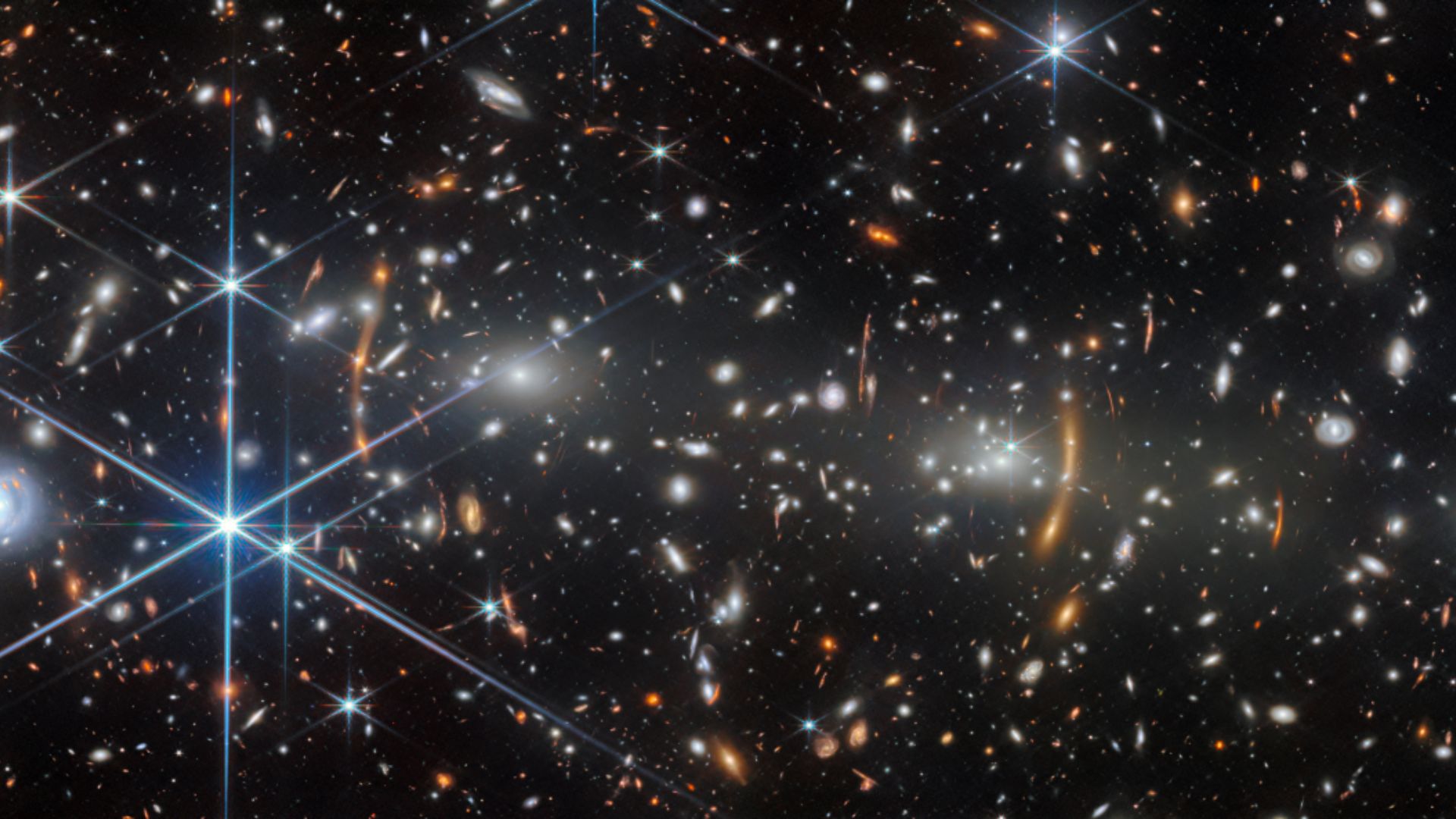 A black space background is speckled by yellow, white, blue and orange stars including blue stars with long diffraction spikes.