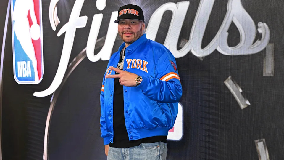 Fat Joe in a blue New York jacket and cap poses in front of an NBA Finals backdrop, making a peace sign.