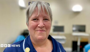 A nurse in a blue uniform with ash blonde hair smiling at the camera standing in a clinical room in a hospital  with computer screens in the background.