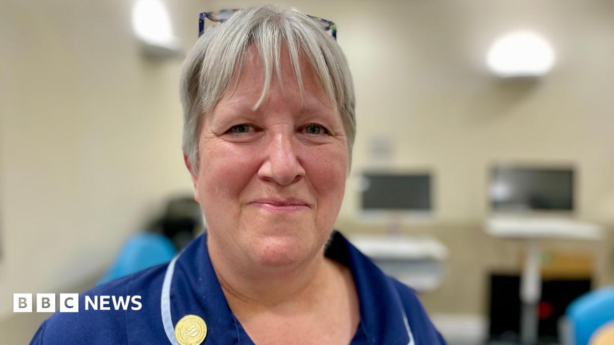 A nurse in a blue uniform with ash blonde hair smiling at the camera standing in a clinical room in a hospital  with computer screens in the background.