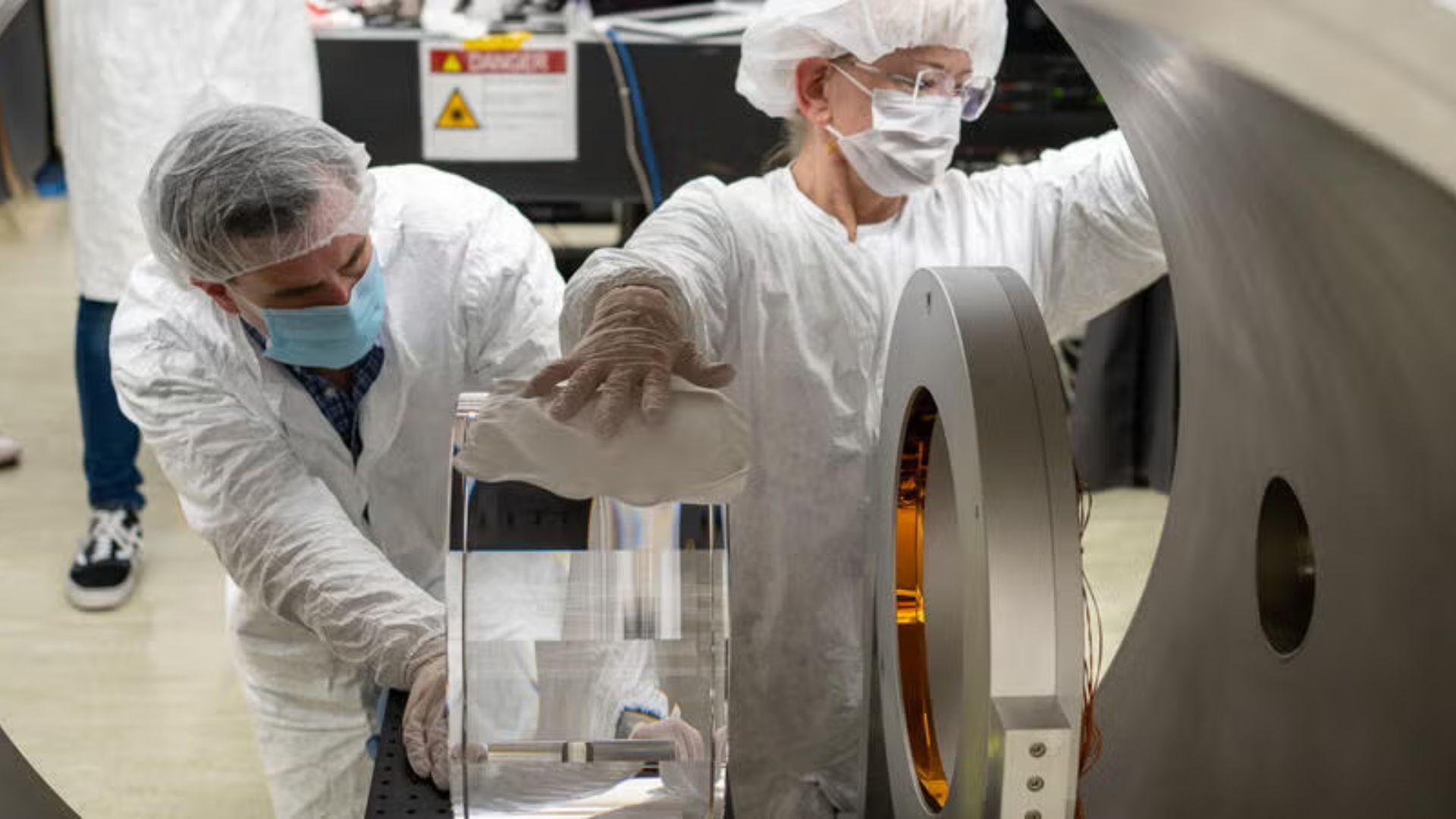 Two people earing white lab coats and other protective lab gear polish a white clear object within a gray cylinder. The view is from within the cylinder.