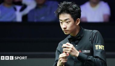Wu Yize, wearing a black shirt and bowtie, chalks his cue with his left hand