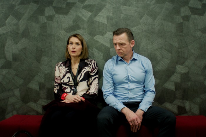 73399_5_S2_Ep5_The Undeclared War - Series 2 Ep5-2 Simon Pegg and Siân Brooke in The Undeclared War, sat next to each other on a bench against a grey wall.