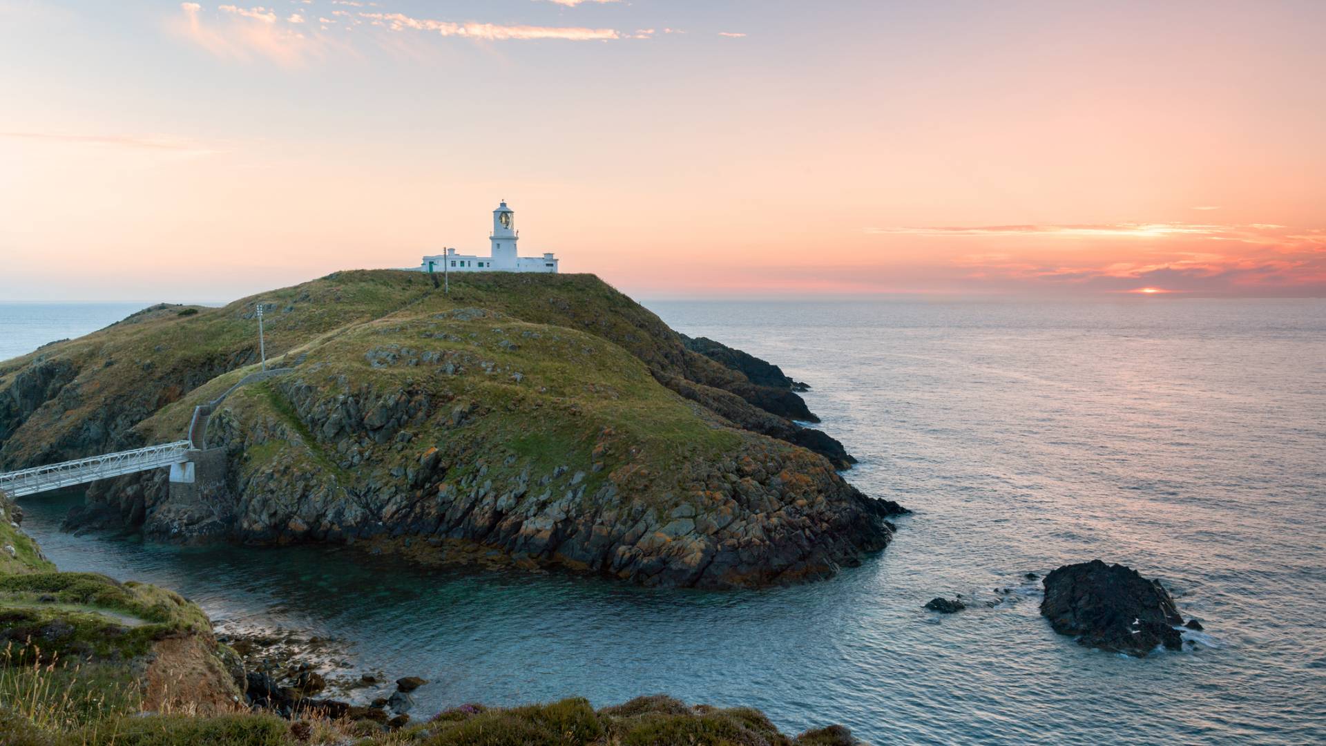 A white lighthouse sits atop a grassy cliff overlooking the calm sea at sunset, with a footbridge connecting the island to the mainland and soft orange and pink hues in the sky.