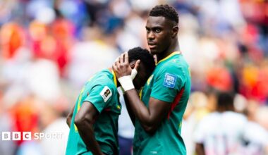 Lamine Camara of Senegal leans his face against the chest of team-mate Moussa Niakhate as he is comforted following the World Cup 2026 last-32 loss to Belgium in Seattle. Niakhate has his hands clasped around the back of Camara's head to pull him in. Both are visible from waist up and wearing their green Senegal jerseys with yellow and red trim. The background is a highly-blurred scene which includes one Belgium player seen from behind and the crowd