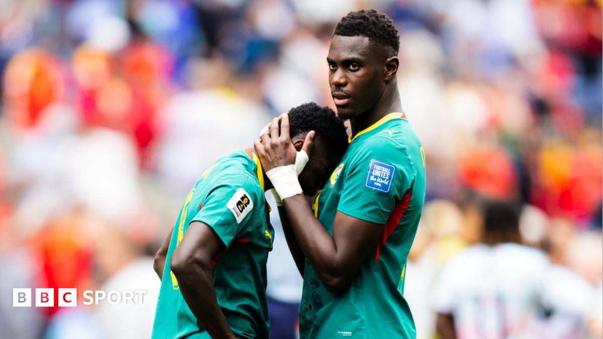 Lamine Camara of Senegal leans his face against the chest of team-mate Moussa Niakhate as he is comforted following the World Cup 2026 last-32 loss to Belgium in Seattle. Niakhate has his hands clasped around the back of Camara's head to pull him in. Both are visible from waist up and wearing their green Senegal jerseys with yellow and red trim. The background is a highly-blurred scene which includes one Belgium player seen from behind and the crowd
