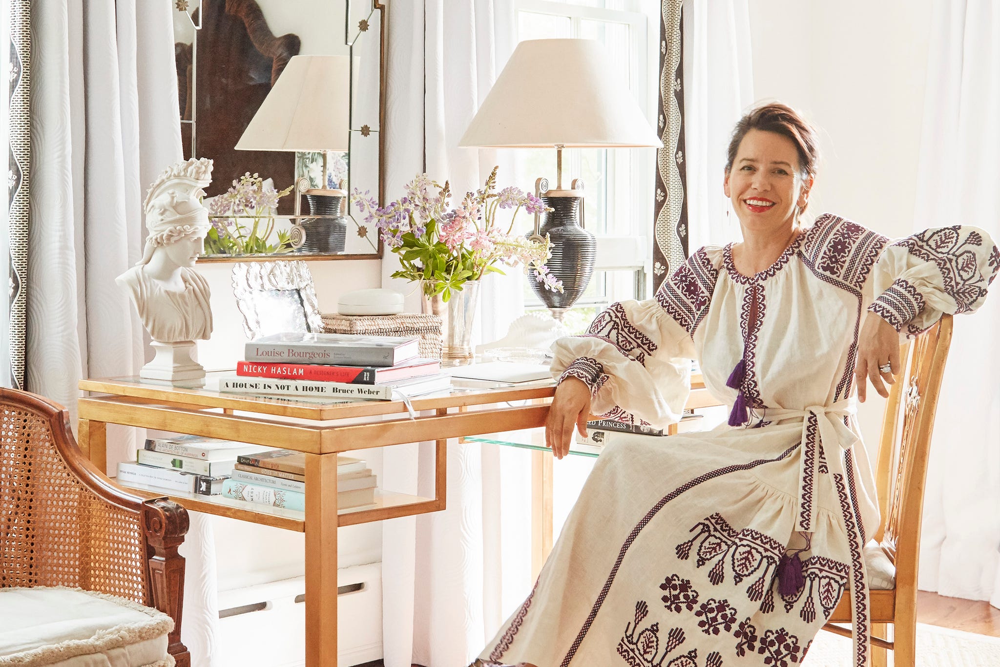 A woman in an embroidered dress sits at a wooden table surrounded by books and flowers in a bright room.