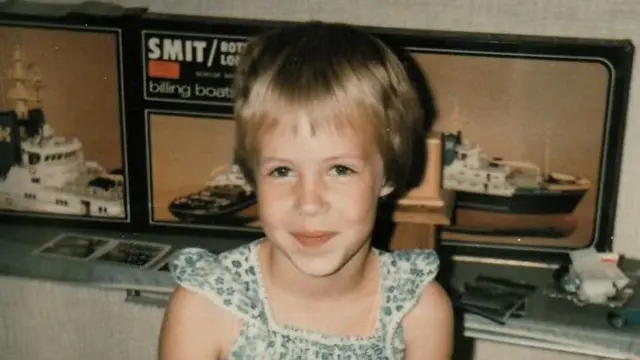 A 1982 photo show Nina Blom at seven years old, inside a house wearing a summer dress. She get short blond hair and get a shy smile on her face. She sidon in front of a display of model ships.