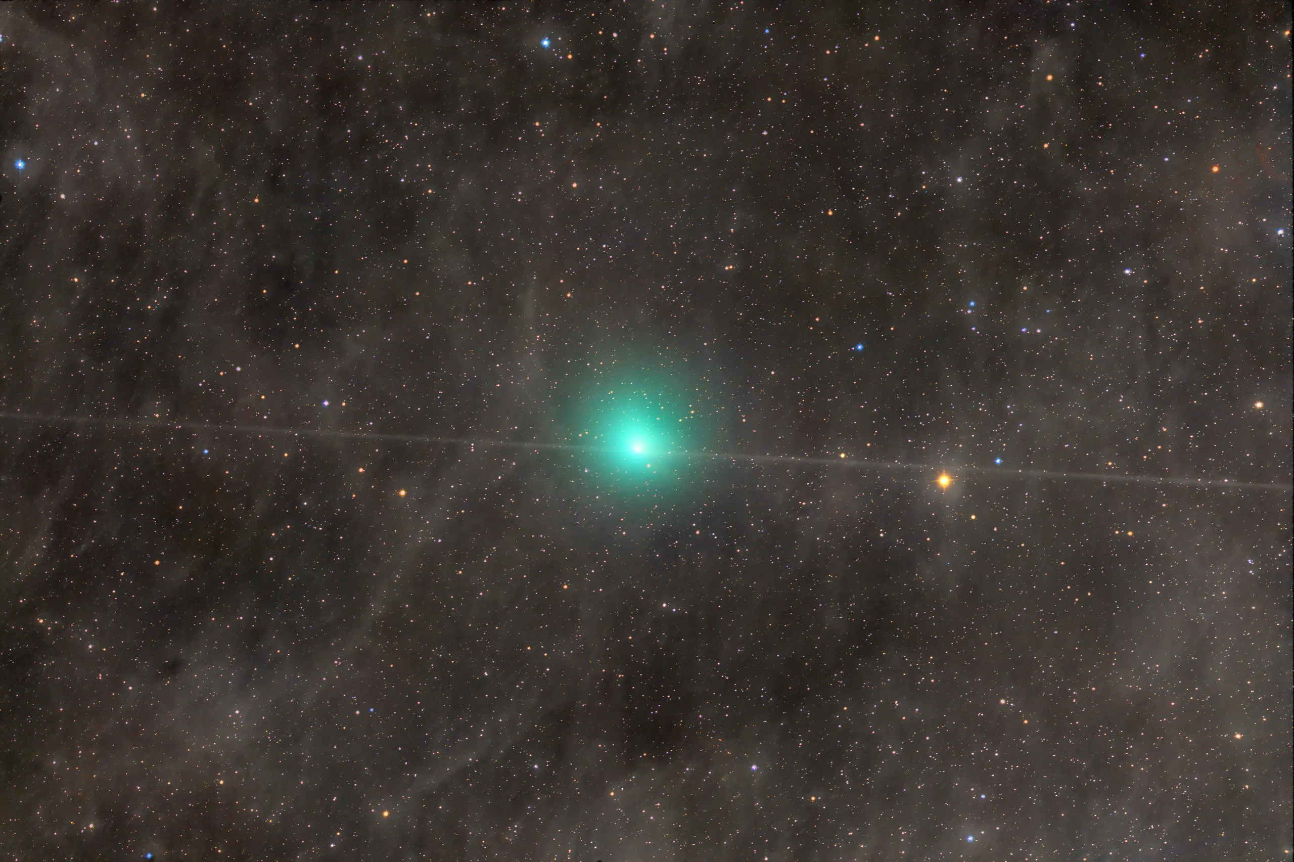 A telescope image of Comet 10P/Tempel 2, showing a bright blue-green coma glowing near the center of a richly star-filled field. A diagonal satellite or aircraft trail crosses through the frame. The surrounding star field is dense with colorful blue, orange, and white stars against a dark background.