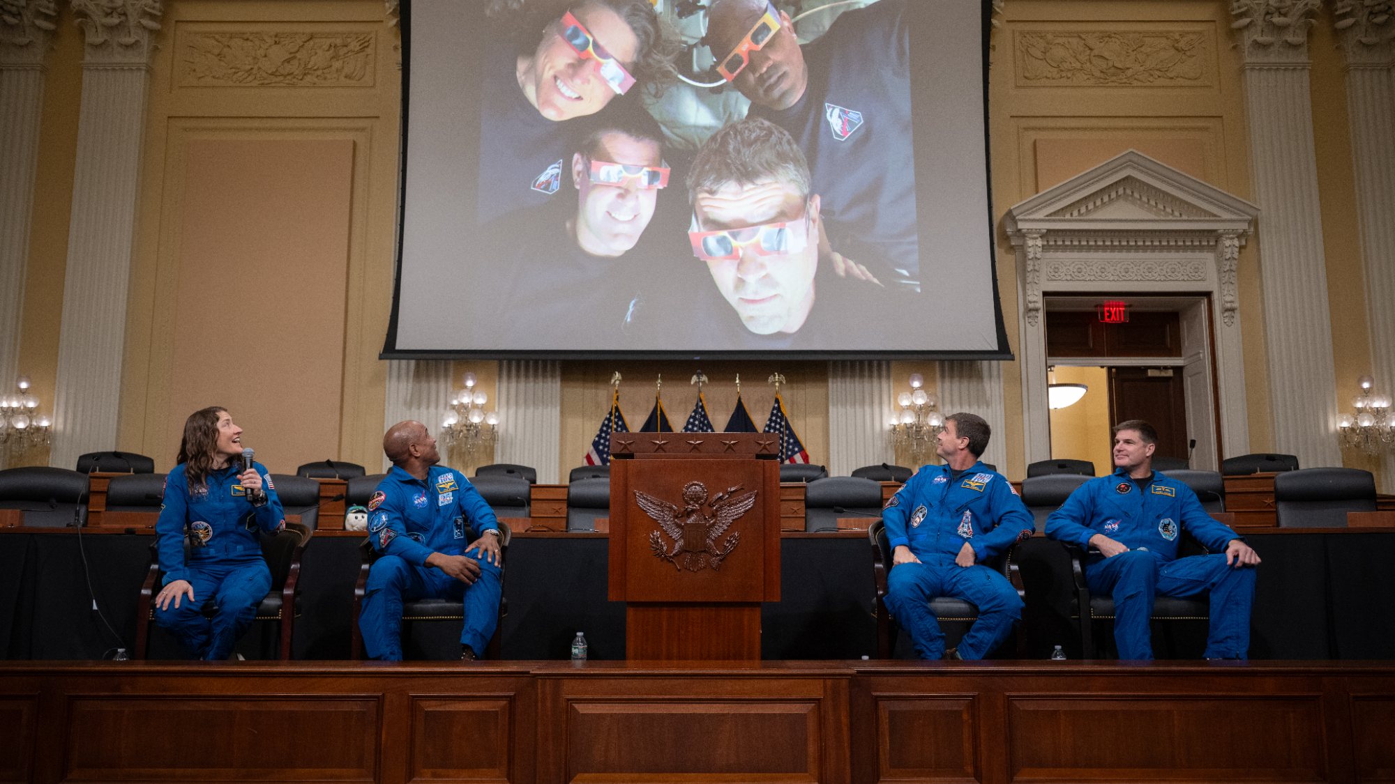 Four Artemis 2 astronauts sit on Capitol Hill and look up at an image from their flight.