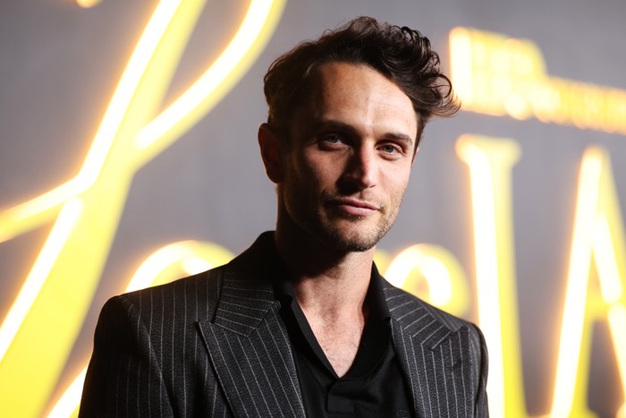 Colin Woodell poses in a black shirt and grey pinstripe blazer in front of a yellow and grey sign