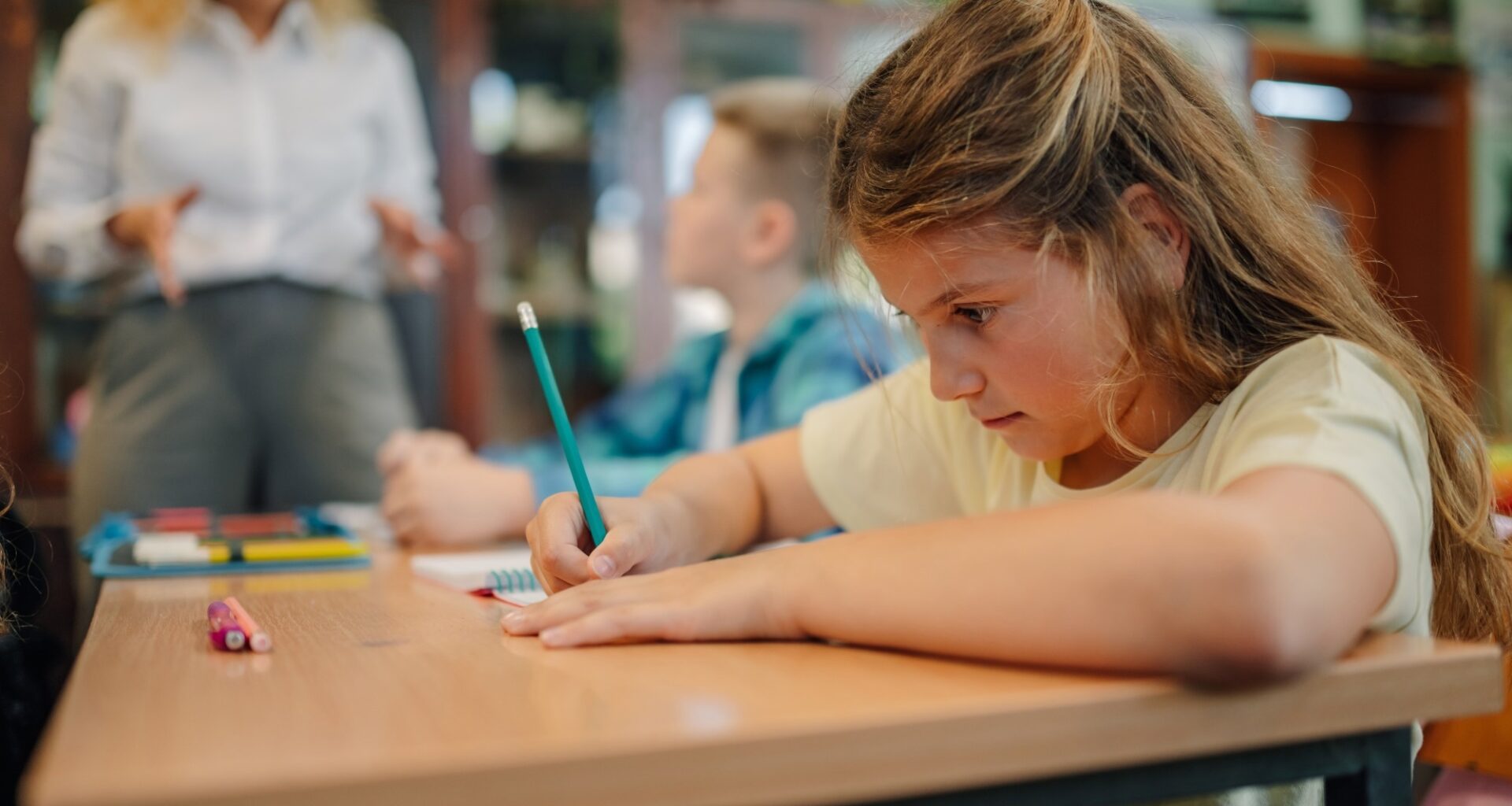 Schoolgirl writing on notebook with pencil during class, while teacher explains lesson to elementary school students