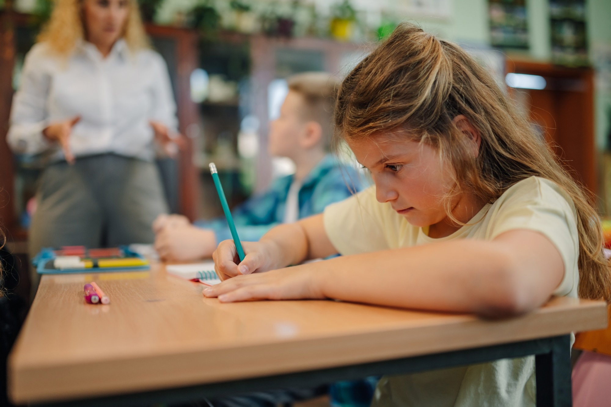 Schoolgirl writing on notebook with pencil during class, while teacher explains lesson to elementary school students