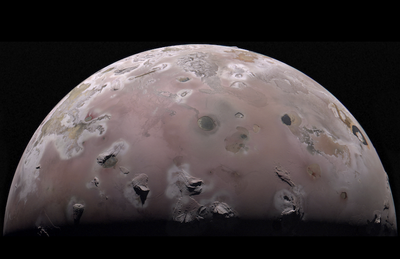 Partial view of brownish planet-like Part of pinkish planet-like sphere pockmarked with bright and dark splotches and tall, isolated peaks.