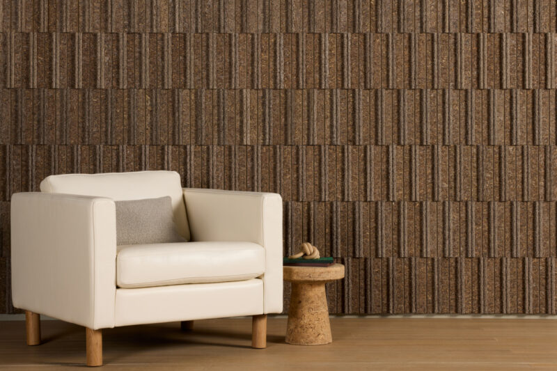 A white armchair with a gray pillow sits next to a small cork side table, against a textured brown wall with a geometric pattern.