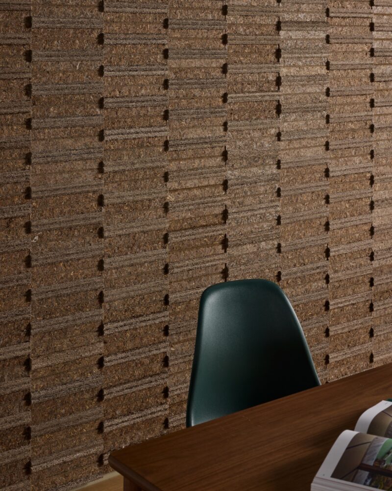 A green chair and wooden table with an open book sit in front of a brown textured wall with horizontal and vertical grooves, accented by eelgrass acoustic wall tiles.