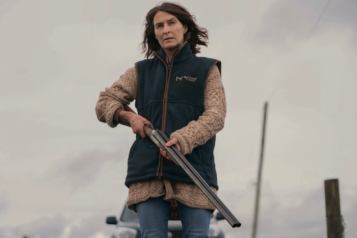 Helen Baxendale as Bethany Morgan standing outside on a grey day and holding a rifle in her hands, looking stressed as she also wears a gilet and shirt.