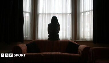 A woman sits on the back of a sofa, looking out of the window in fear of her partner, coming home after football.