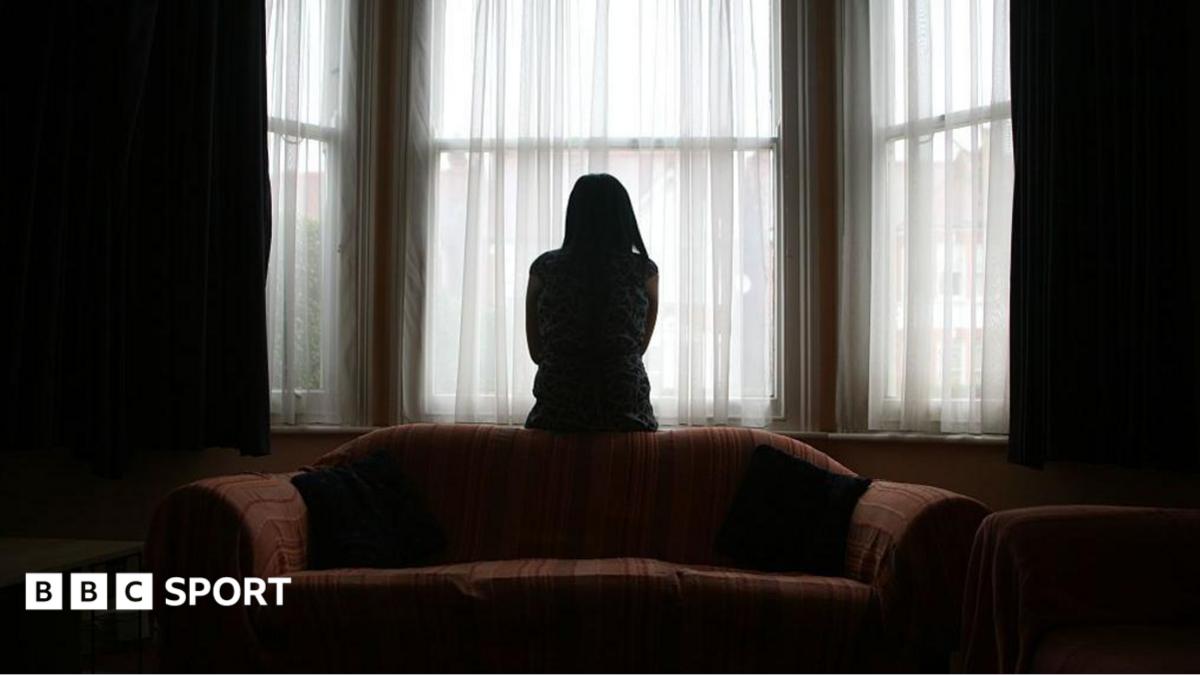 A woman sits on the back of a sofa, looking out of the window in fear of her partner, coming home after football.