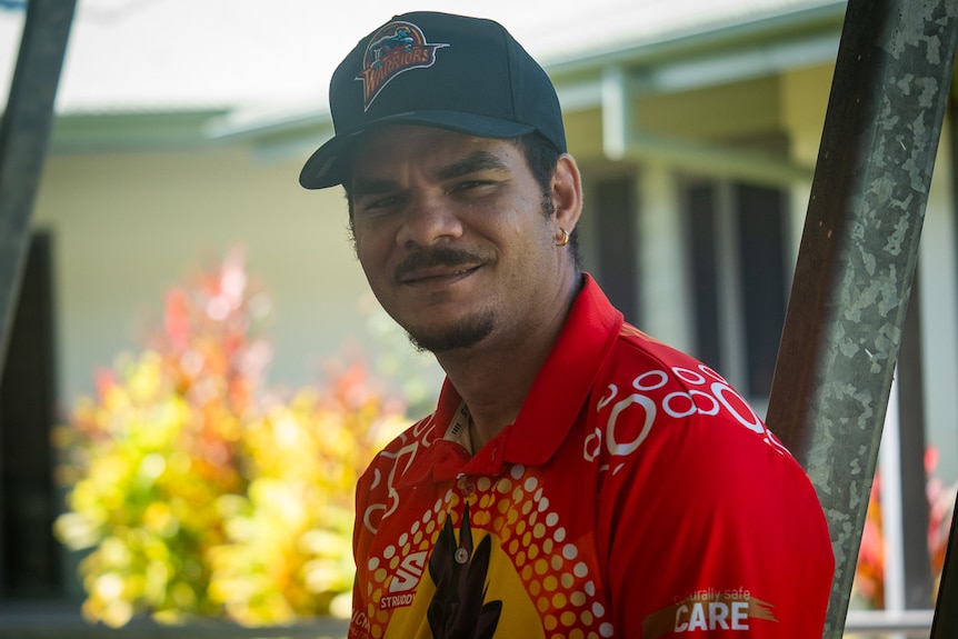 A man in a red t-shirt with Aboriginal art designs and a cap looks towards the camera with a neutral expression.