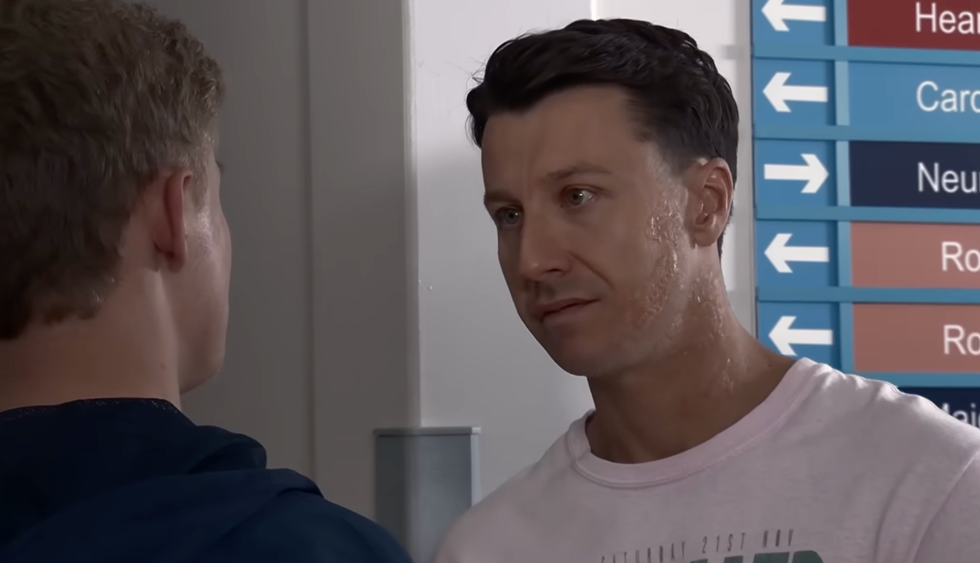 two men talking in a hospital corridor next to directional signs.