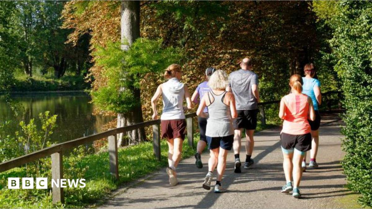 A group of people going for a run in parkland. There is a body of water to the left of the image, with trees and a small wooden fence