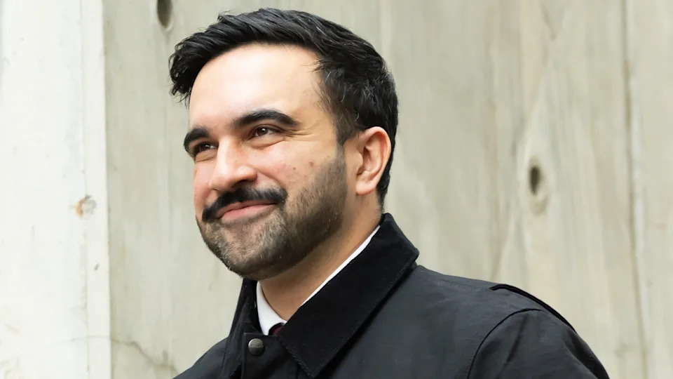 Zohran Mamdani with dark hair and a beard, smiling, wearing a dark coat, standing against a light-colored wall.