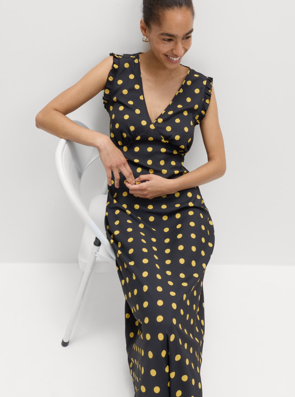 person in a black dress with yellow polka dots sitting on a white chair, smiling.