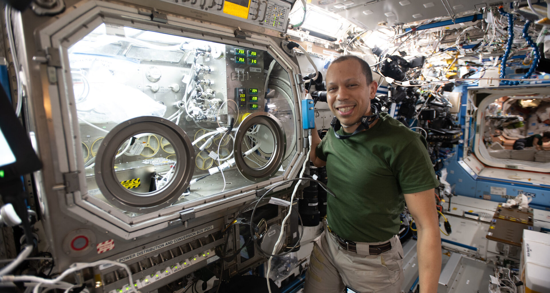 NASA astronaut Chris Williams shows off the Destiny laboratory module’s Microgravity Science Glovebox aboard the International Space Station. WIlliams was supporting semiconductor crystal research to help advance the commercial space economy and promoting Earth-based industries. He is wearing a dark green t-shirt and khaki pants with a brown belt. He is shown smiling with a headset around his neck.