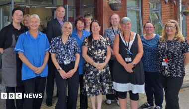 Staff members of the care home stand for a photo, they are smiling. Some wear nursing scrubs, while others wear office attire.