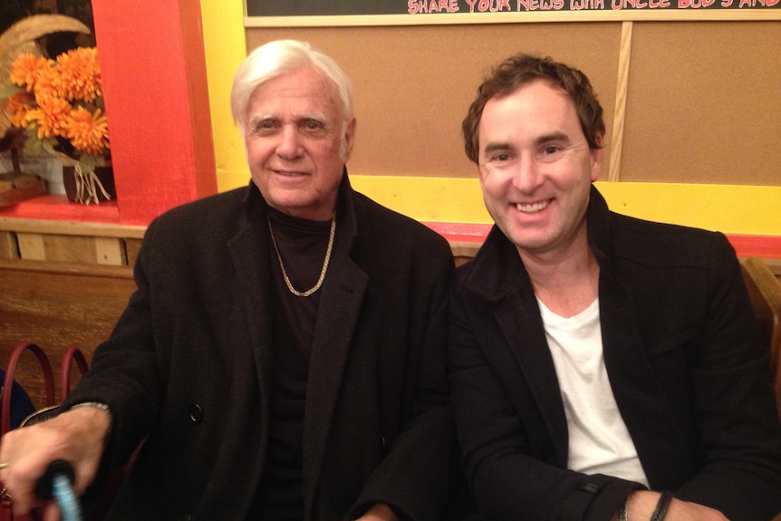 Joe Melson wears a black coat and black shirt sitting next to Damien in a black jacket and white shirt.