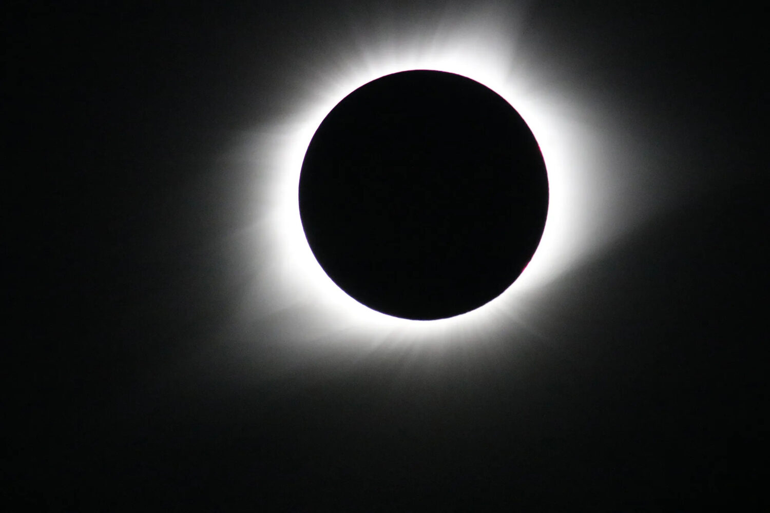 A stark, high-contrast photograph captures a solid black circle perfectly centered against a jet-black background. A bright, glowing ring of white light radiates outward from behind the dark circle, creating a soft, hazy halo with faint streaks extending into the surrounding darkness.