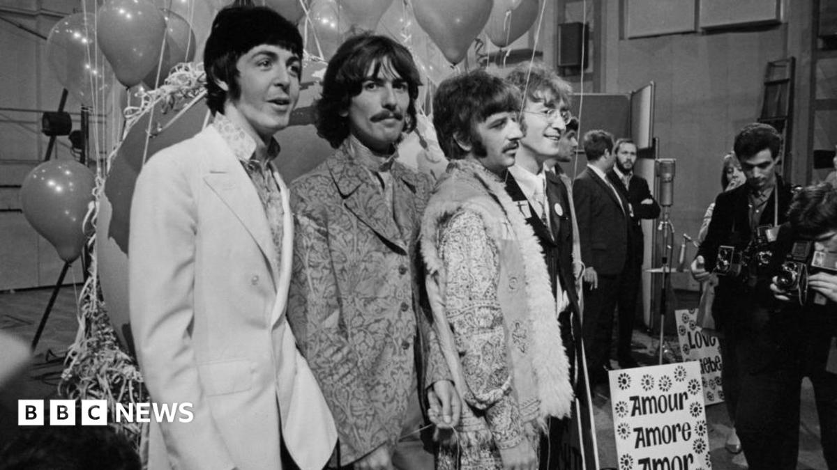 The Beatles post for a photo. Their clothing is from 1967. The photo is black and white. Some photographers are visible in the side of the image.
