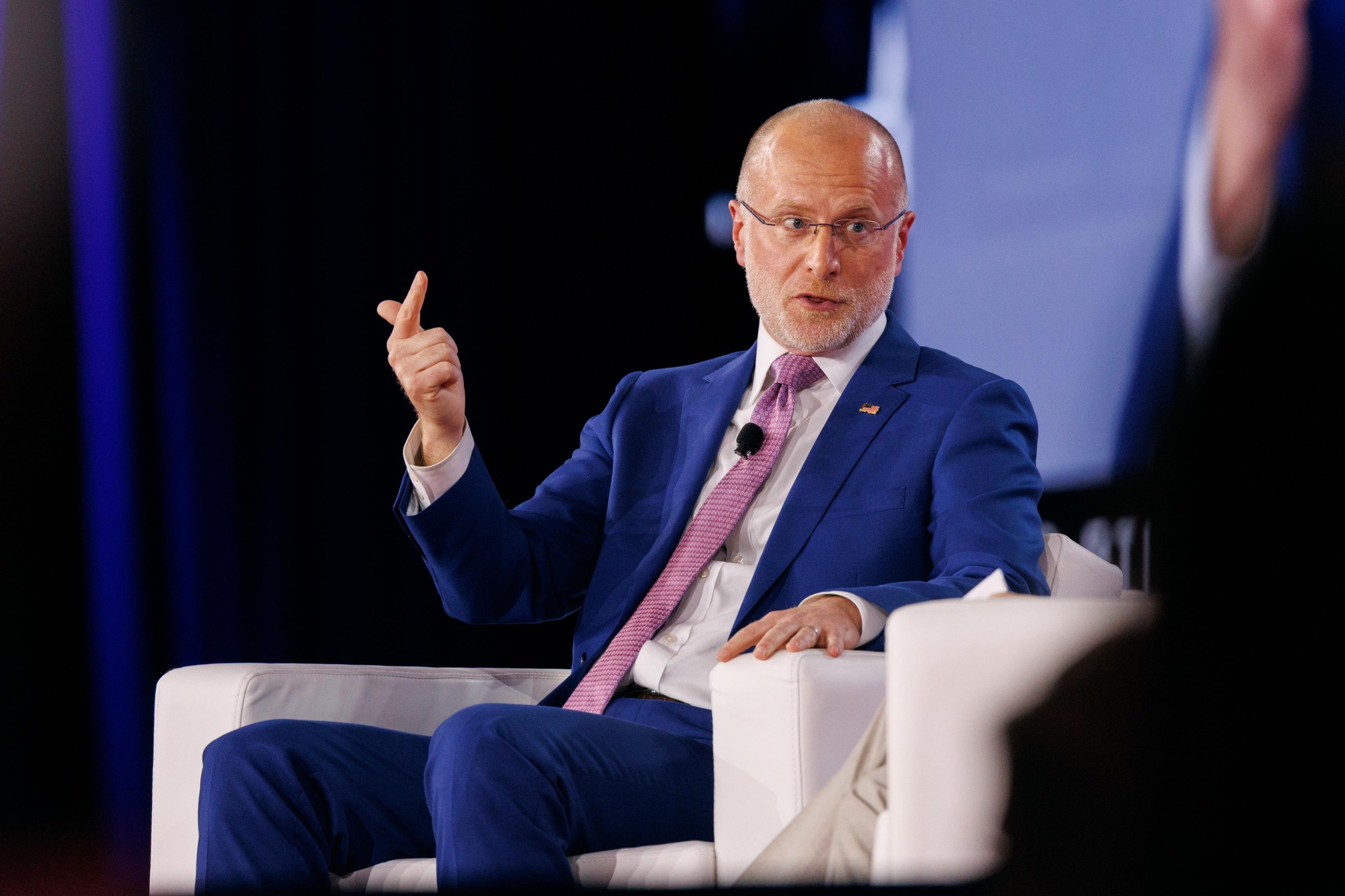 Brendan Carr, in a blue suit, speaks on stage while sitting in a white chair.