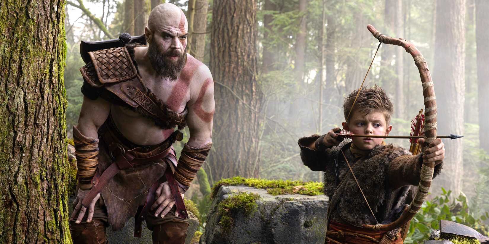 Kratos leaning over Atreus drawing a bow in God of War.