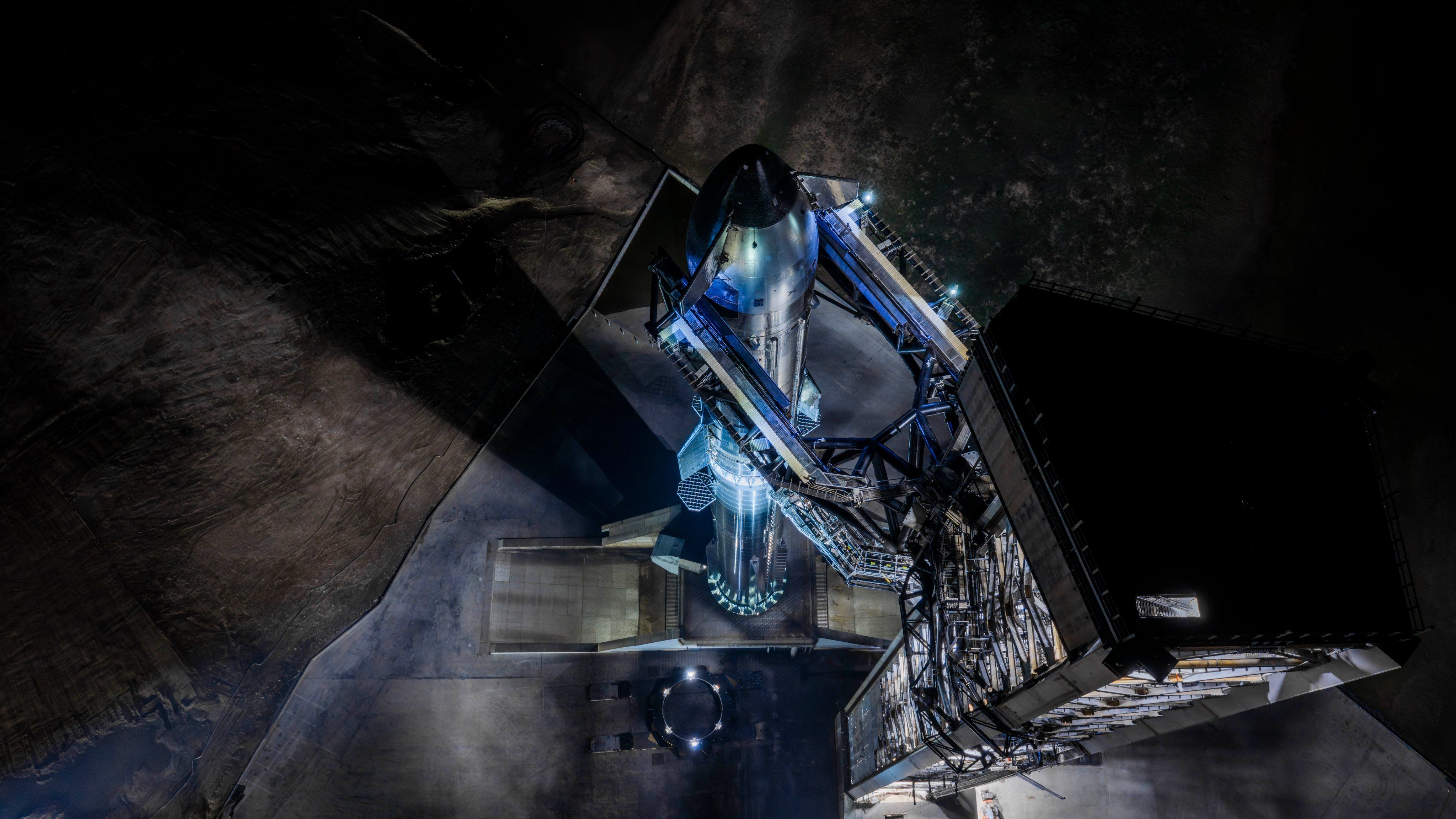 A giant steel rocket stands next to a launch tower lit by spotlights in the dark of night.