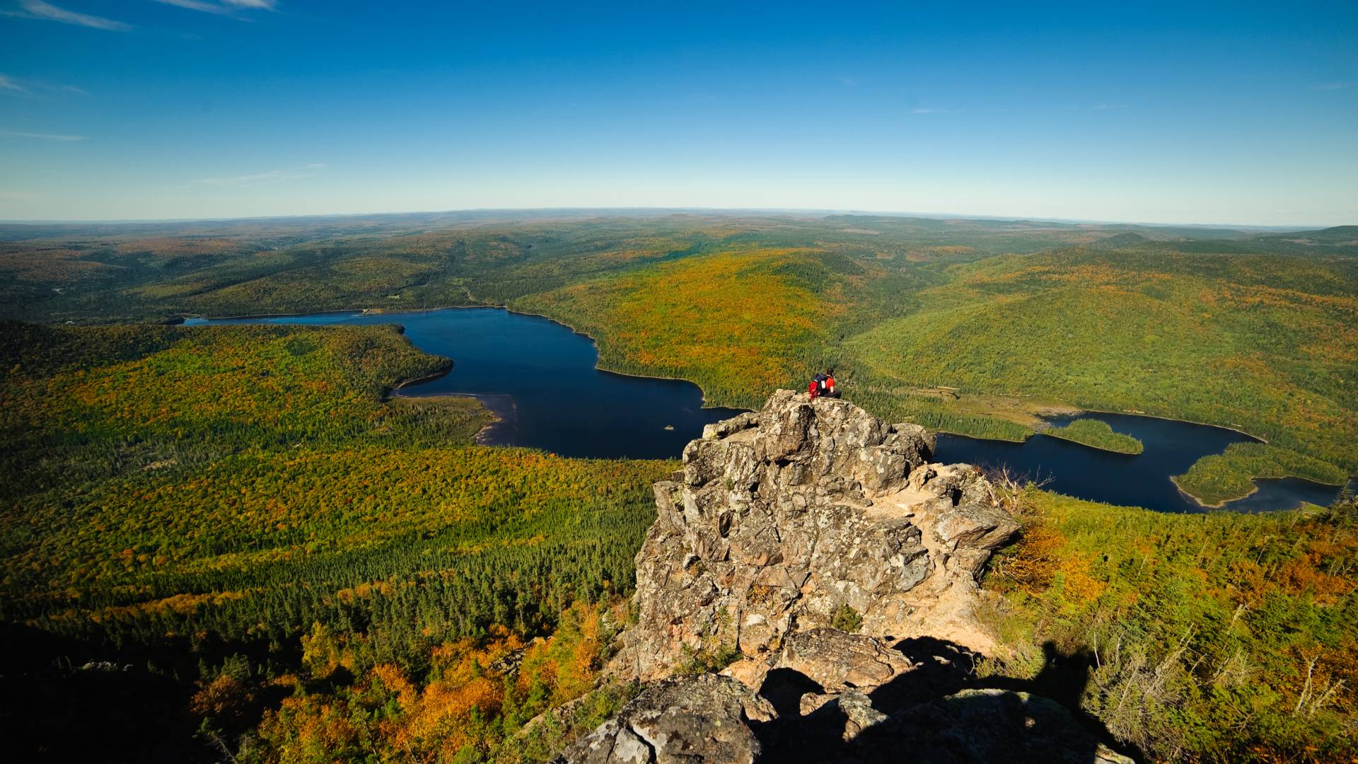 A rocky cliff overlooks a vast landscape of green and orange forests, with winding blue lakes below under a clear blue sky. Two people in red sit at the edge of the cliff enjoying the scenic view.
