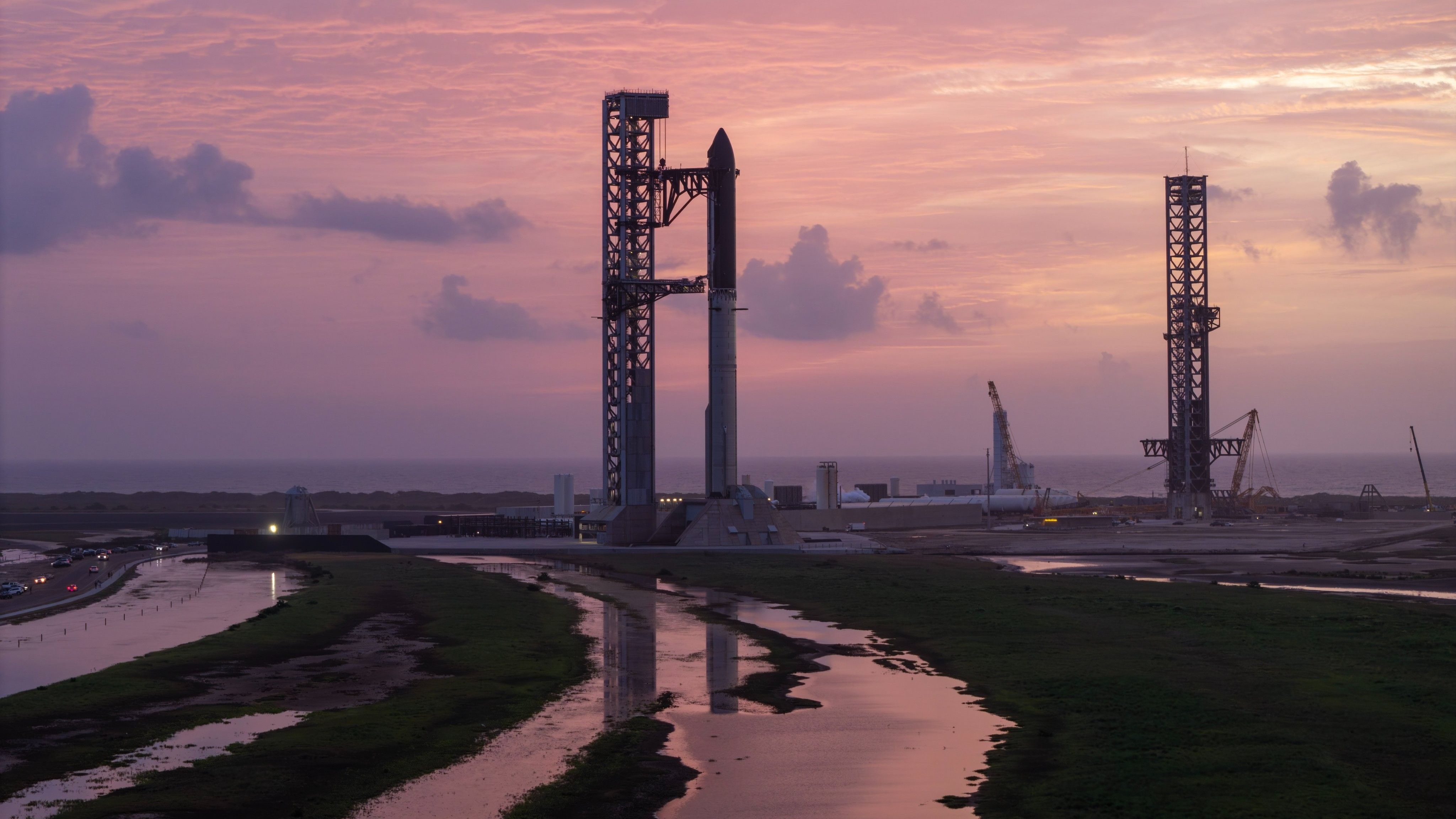 A giant steel rocket stands next to a launch tower reflected in streams of water in the foreground against a rich pink cloudy sky