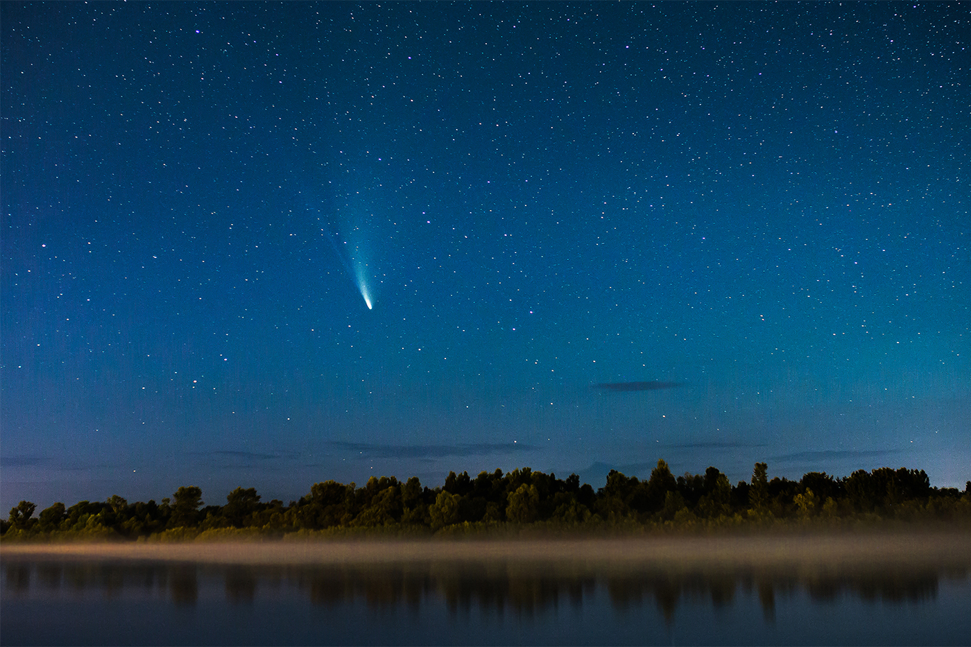 A bright-tailed comet streaks across a starry night sky above a fog-covered river and treeline.