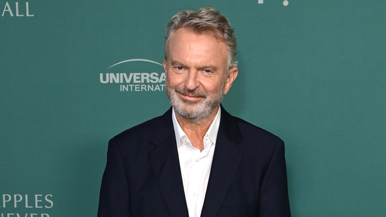 Sam Neill with gray hair and a gray beard wearing a dark suit and a white shirt with an open collar.