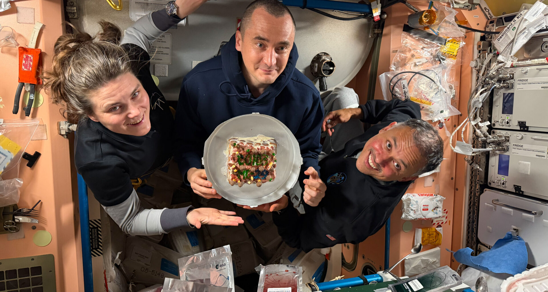 From left, Expedition 74 flight engineers Anna Kikina and Pyotr Dubrov of Roscosmos, and Anil Menon of NASA pose for a portrait while holding a cake celebrating their recent arrival aboard the International Space Station.