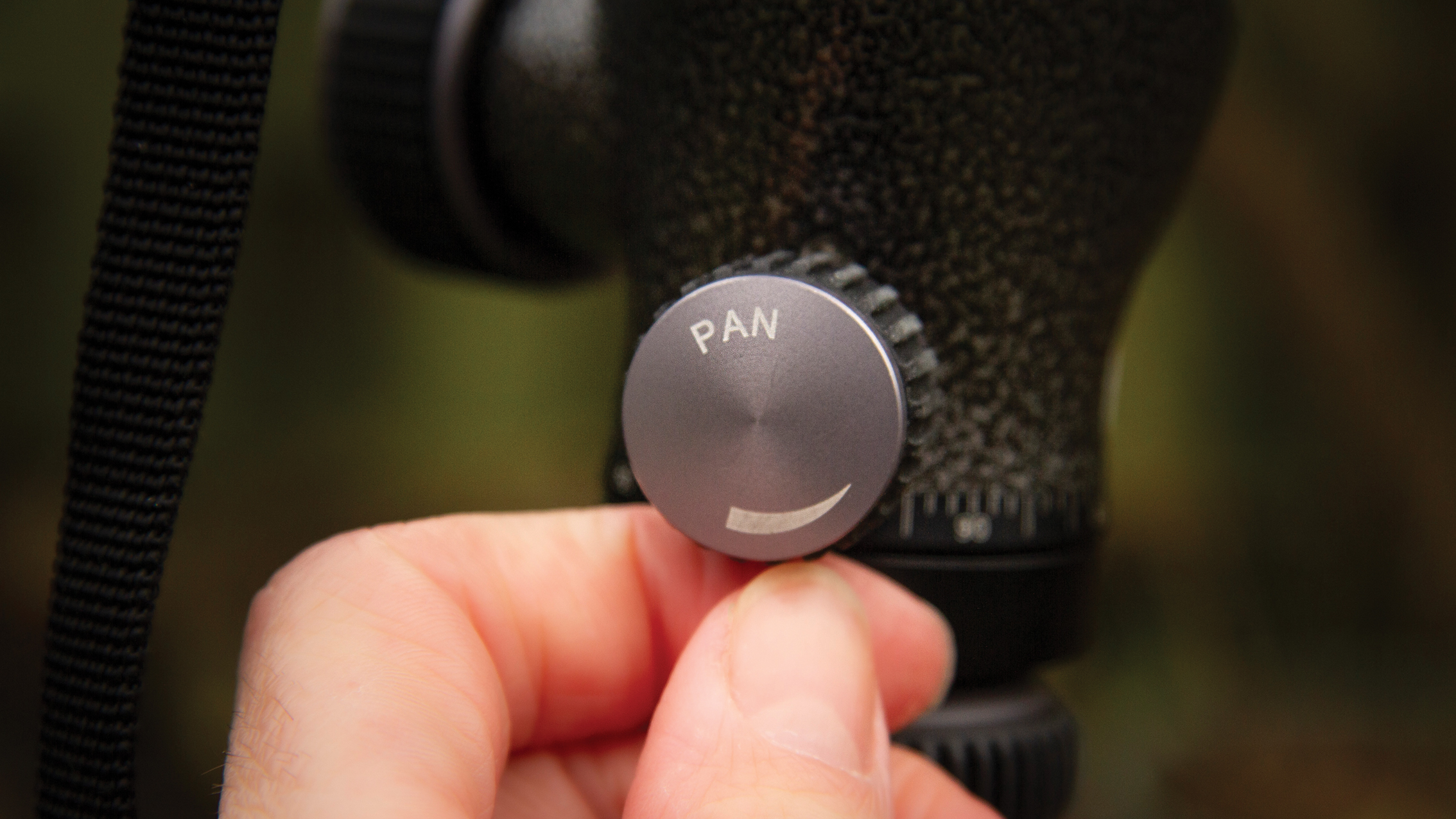 Hand adjusting the "PAN" knob on a black textured tripod or camera mount with blurred green-brown background