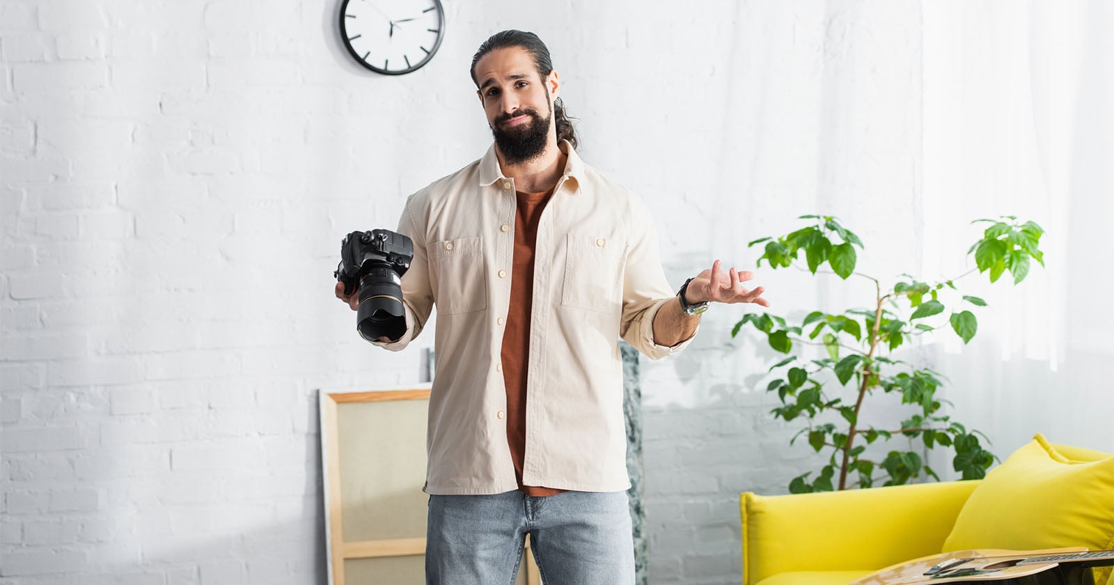 A man with a beard holds a camera in one hand and shrugs with the other, standing in a bright room with a yellow couch, green plant, and a clock on the wall.