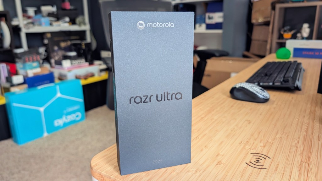 Box of the Motorola Razr Ultra smartphone displayed on a wooden surface with various items in the background.
