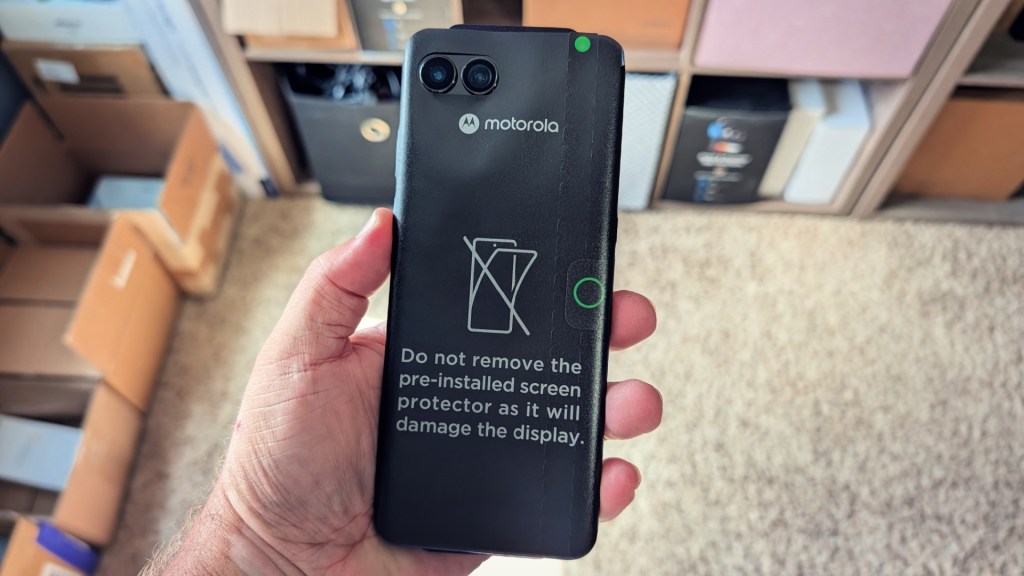 A person holding a Motorola smartphone with a warning message about not removing the pre-installed screen protector, set against a background of cardboard boxes.