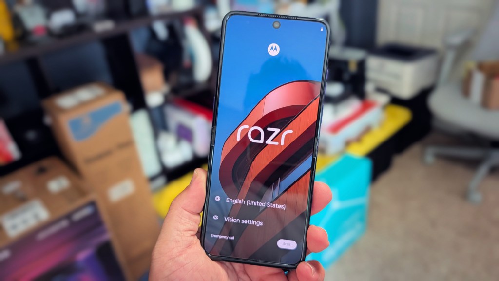 A person holding a Motorola Razr smartphone displaying the setup screen with options for language and vision settings. Background features blurred boxes and electronic items.