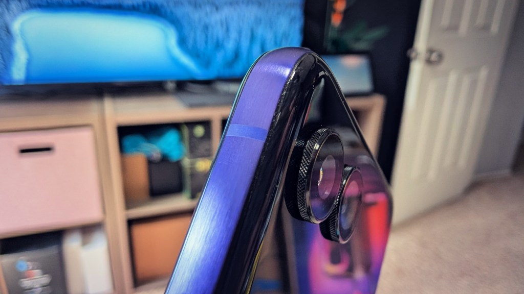 Close-up view of a smartphone with a dual camera setup, showing the edge and back of the device, with a blurred background featuring a television and storage units.