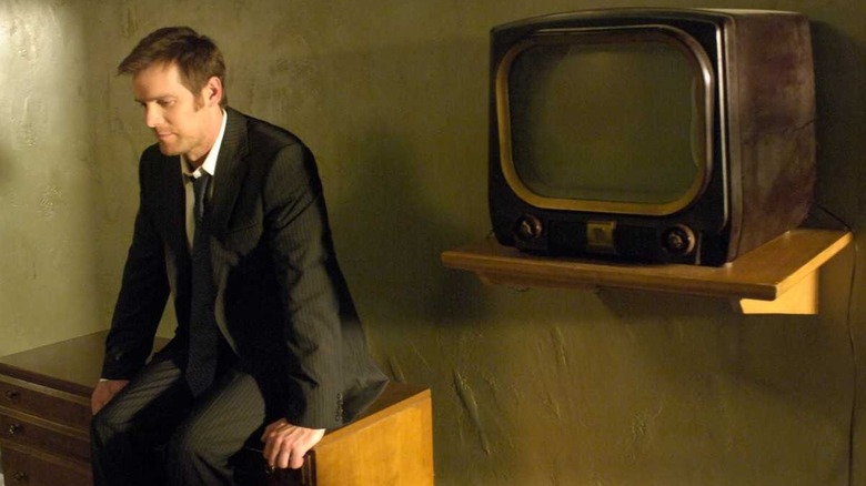 Peter Krause as the detective Joe Miller, sitting on a chest of drawers next to an old 1960s television set, on Syfy's "The Lost Room."