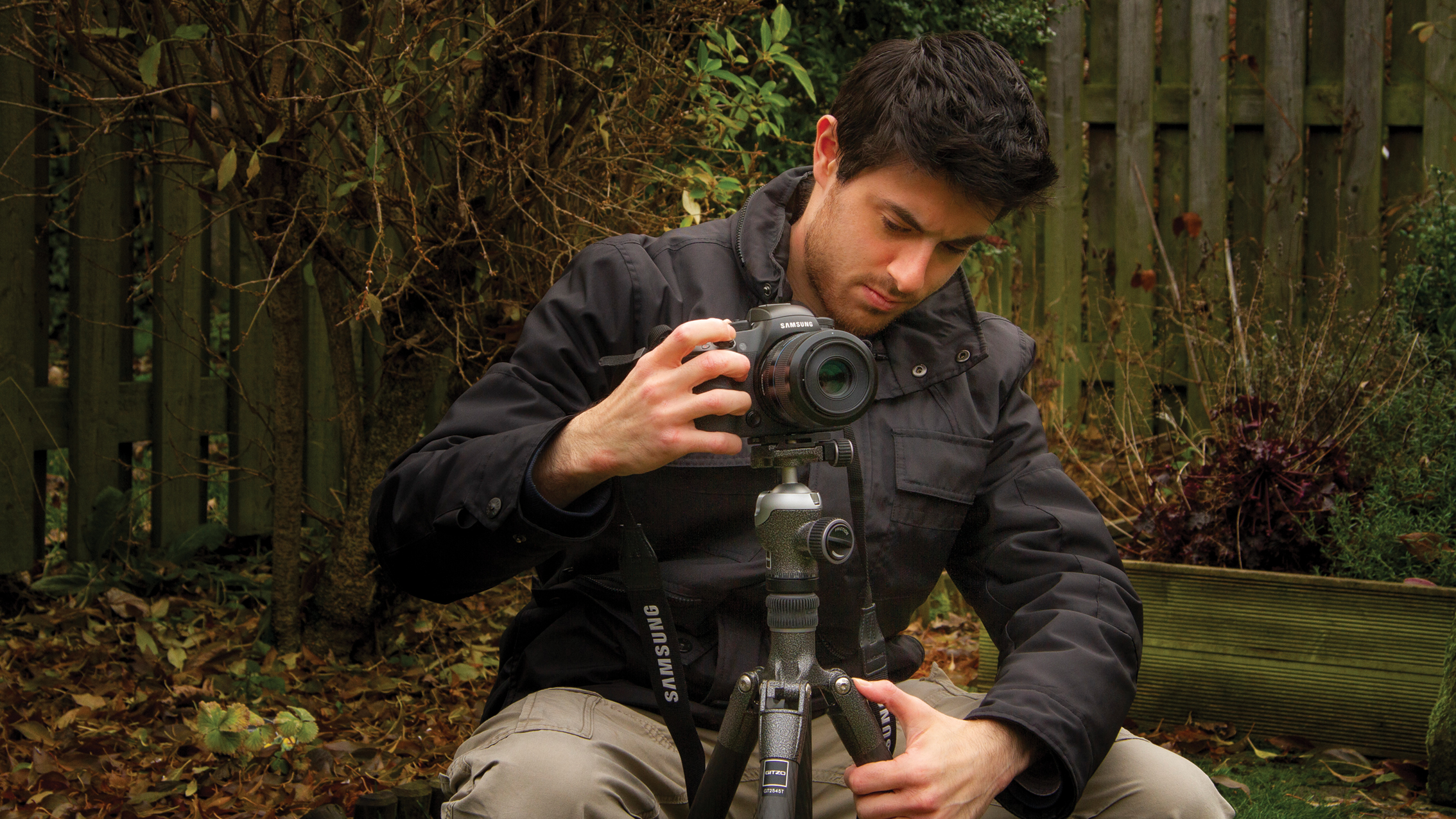 Man adjusting a DSLR camera on a tripod in a garden with dry leaves and a wooden fence background